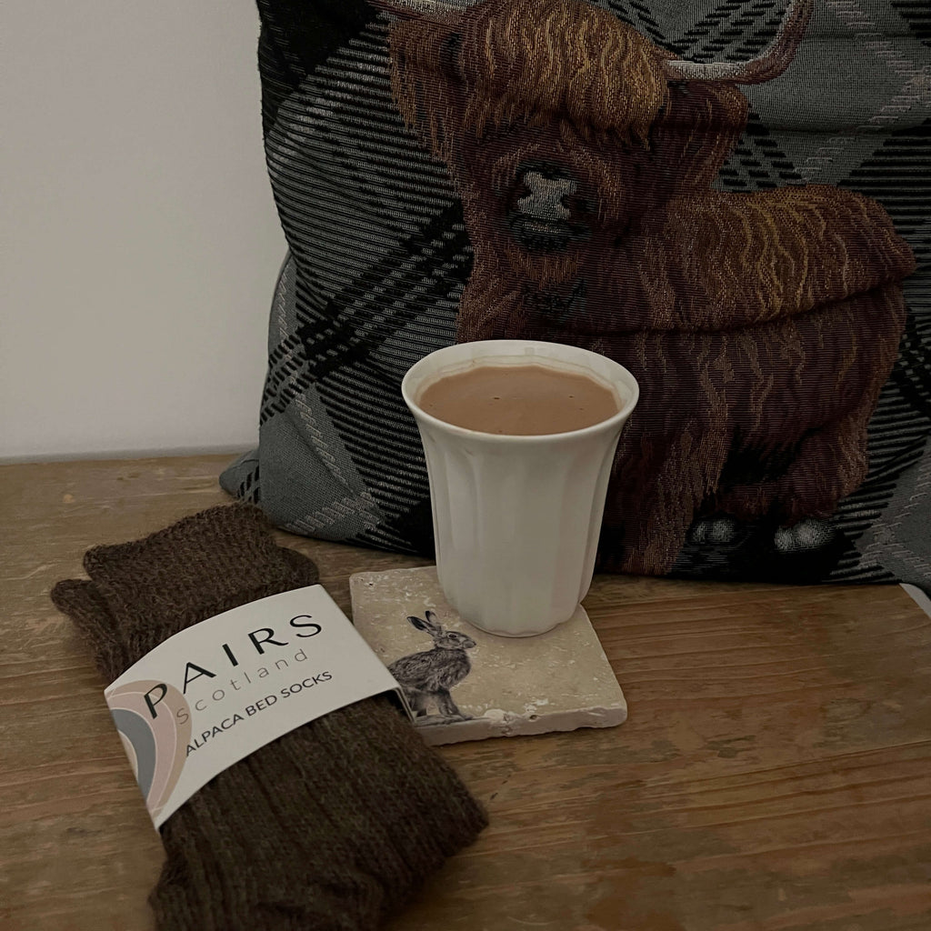 Pair of brown alpaca bed socks with a label, white mug with hot chocolate, and highland cow cushion.
